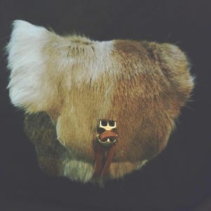 May include: A small handbag crafted from textured fur in shades of white, tan, and brown. The bag features a front flap closure secured by a gold-toned metal buckle and a thin brown leather strap that hangs vertically from the center of the clasp.