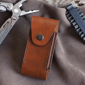 May include: A brown leather multi-tool sheath with a snap closure, a silver multi-tool, and a black bit kit. The multi-tool is open, displaying pliers and a knife. The bit kit and multi-tool are both branded with the name "LEATHERMAN".