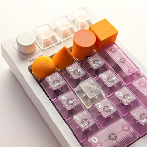 May include: A white mechanical keyboard with clear and pink keycaps. The keyboard features a variety of orange geometric shapes and clear keycaps. The keys are labeled with numbers and symbols.
