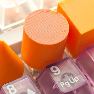 May include: Close-up of a mechanical keyboard with orange and purple keycaps. The orange keycaps are cylindrical and square, while the purple keycaps have white lettering and numbers. The keyboard is a tech accessory.