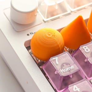 May include: Close-up of a white mechanical keyboard with clear and pink keycaps. The keyboard features an orange spherical key and an orange triangular key. A white cylindrical knob is also visible.