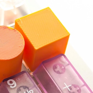 May include: Close-up of colorful keyboard keys. The image features a bright orange cube and cylinder, along with translucent purple keys with white symbols and text. The keys are arranged against a white background, creating a vibrant and modern aesthetic.