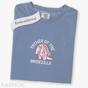 May include: A blue t-shirt featuring the embroidered text "FATHER OF THE BRIDEZILLA" and a red and white dinosaur design. The shirt has a tag that says "Embroidered".
