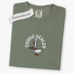 Op de afbeelding: Een olijfgroen t-shirt met ronde hals en een geborduurde afbeelding van een witte gans met een koksmuts die een dienblad met koekjes vasthoudt. De tekst "COOKIE DEALER" staat in witte letters in een boog boven de gans geborduurd.