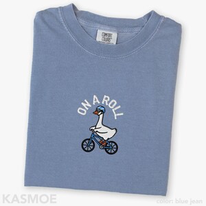 May include: A blue jean colored t-shirt featuring an embroidered graphic of a white goose wearing a blue helmet and orange shoes while riding a blue bicycle. The text ON A ROLL is embroidered in white above the goose. The shirt has a Comfort Colors label.