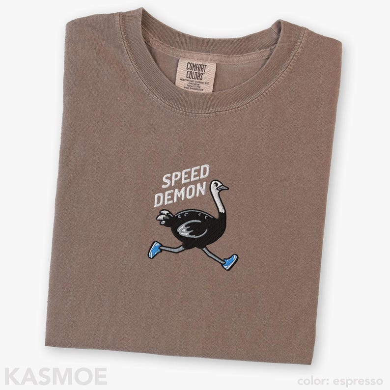 May include: An espresso-colored t-shirt featuring an embroidered graphic of a black ostrich running while wearing blue sneakers. Above the ostrich, the text SPEED DEMON is embroidered in white. The shirt has a crew neckline and a Comfort Colors brand label.