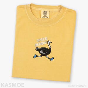 May include: A mustard yellow t-shirt featuring an embroidered graphic of a black ostrich running while wearing blue sneakers. Above the ostrich, the text SPEED DEMON is embroidered in white. The shirt has a Comfort Colors brand label at the neck.