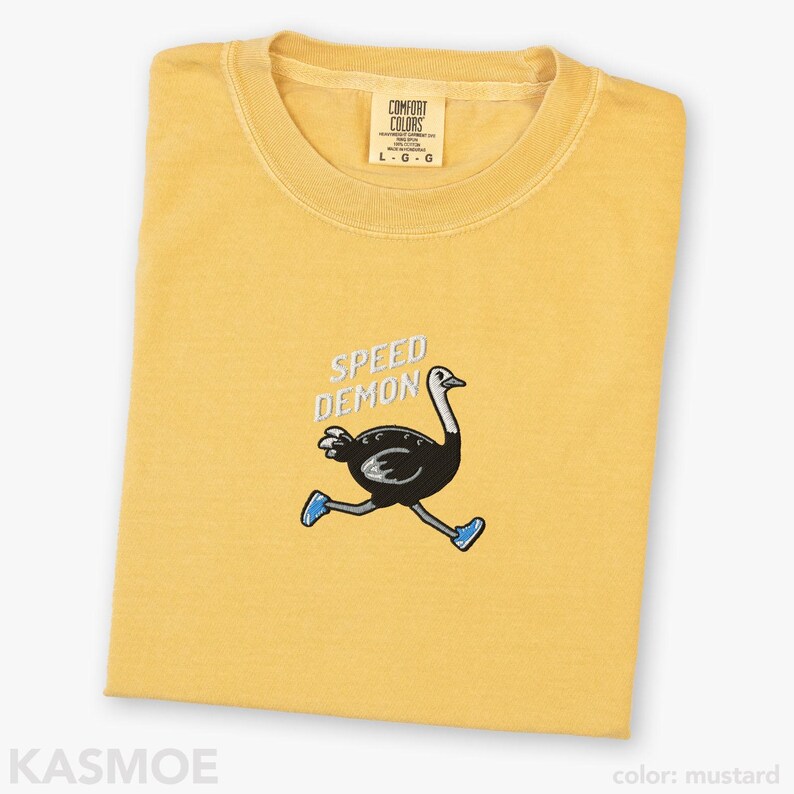 May include: A mustard yellow t-shirt featuring an embroidered graphic of a black ostrich running while wearing blue sneakers. Above the ostrich, the text SPEED DEMON is embroidered in white. The shirt has a Comfort Colors brand label at the neck.