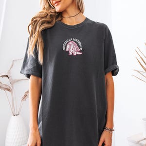 Może przedstawiać: Ciemnoszary t-shirt z kr&oacute;tkim rękawem z małym haftowanym wzorem na piersi. Grafika przedstawia r&oacute;żowego dinozaura w białym welonie ślubnym z napisem BRIDEZILLA WRANGLER ułożonym w łuk wok&oacute;ł postaci.