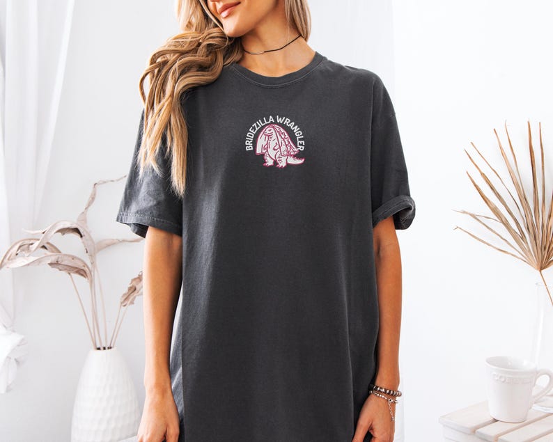 Może przedstawiać: Ciemnoszary t-shirt z kr&oacute;tkim rękawem z małym haftowanym wzorem na piersi. Grafika przedstawia r&oacute;żowego dinozaura w białym welonie ślubnym z napisem BRIDEZILLA WRANGLER ułożonym w łuk wok&oacute;ł postaci.