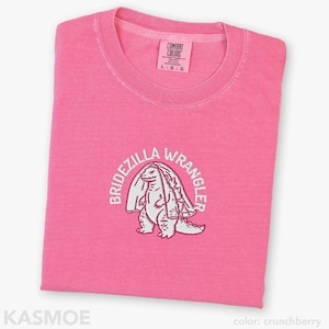 Może przedstawiać: Jaskrawor&oacute;żowy t-shirt z wyhaftowaną białą grafiką przedstawiającą stworzenie w welonie ślubnym. Tekst "BRIDEZILLA WRANGLER" jest wygięty w łuk nad stworzeniem. Wewnętrzna metka na kołnierzu zawiera napis "COMFORT COLORS" oraz rozmiar "L".