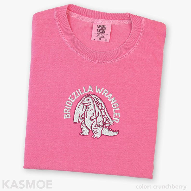 Może przedstawiać: Jaskrawor&oacute;żowy t-shirt z wyhaftowaną białą grafiką przedstawiającą stworzenie w welonie ślubnym. Tekst "BRIDEZILLA WRANGLER" jest wygięty w łuk nad stworzeniem. Wewnętrzna metka na kołnierzu zawiera napis "COMFORT COLORS" oraz rozmiar "L".