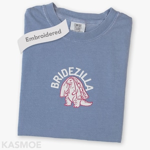 Puede incluir: Una camiseta azul con la palabra "Embroidered" en una etiqueta blanca. La camiseta presenta un diseño bordado en blanco y rojo de un dinosaurio con velo, con la palabra "BRIDEZILLA" encima. La marca "KASMOE" está en la parte inferior.