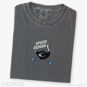 May include: A charcoal gray t-shirt featuring an embroidered graphic of a black ostrich running while wearing blue sneakers. Above the ostrich, the text SPEED DEMON is embroidered in white. The shirt has a crew neckline and a Comfort Colors brand label.