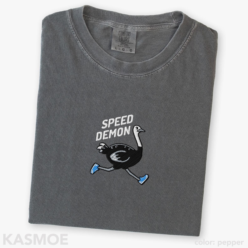 May include: A charcoal gray t-shirt featuring an embroidered graphic of a black ostrich running while wearing blue sneakers. Above the ostrich, the text SPEED DEMON is embroidered in white. The shirt has a crew neckline and a Comfort Colors brand label.