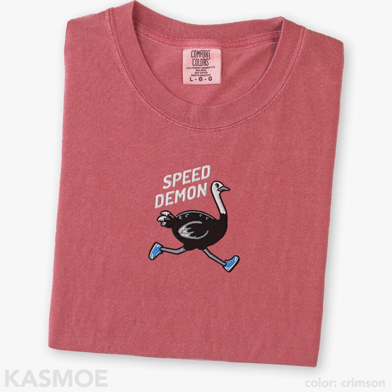 May include: A crimson short-sleeved t-shirt featuring an embroidered graphic of a black ostrich running while wearing blue sneakers. Above the ostrich, the text reads SPEED DEMON in white. The shirt has a Comfort Colors brand label at the neck.