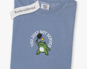 Puede incluir: Una camiseta azul de cuello redondo con un bordado de una rana verde sosteniendo una raqueta de tenis negra y una pelota de tenis amarilla. El texto "YOU JUST GOT SERVED" aparece arqueado sobre la rana. Una etiqueta blanca dice "Embroidered".