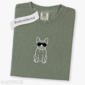 May include: An olive green t-shirt featuring a white embroidered outline of a French bulldog wearing black sunglasses. The shirt has a crew neckline and a Comfort Colors brand label at the inner collar with the text L G G visible.