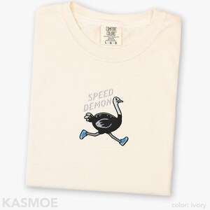 May include: An ivory t-shirt featuring an embroidered graphic of a black ostrich running while wearing blue sneakers. Above the ostrich, the text SPEED DEMON is embroidered in light blue. The shirt has a crew neckline and a Comfort Colors brand label.