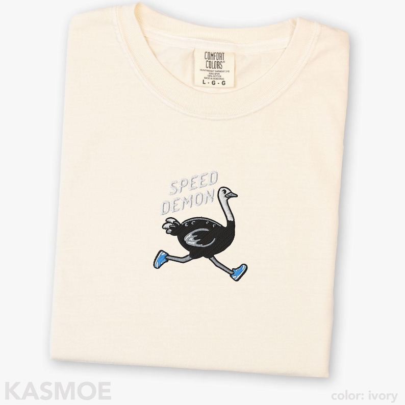 May include: An ivory t-shirt featuring an embroidered graphic of a black ostrich running while wearing blue sneakers. Above the ostrich, the text SPEED DEMON is embroidered in light blue. The shirt has a crew neckline and a Comfort Colors brand label.