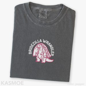 Może przedstawiać: Ciemnoszary t-shirt z wyhaftowaną grafiką przedstawiającą stworzenie przypominające dinozaura w welonie ślubnym. Napis BRIDEZILLA WRANGLER jest wygięty w łuk nad stworzeniem białymi wielkimi literami. Koszulka posiada okrągły dekolt.