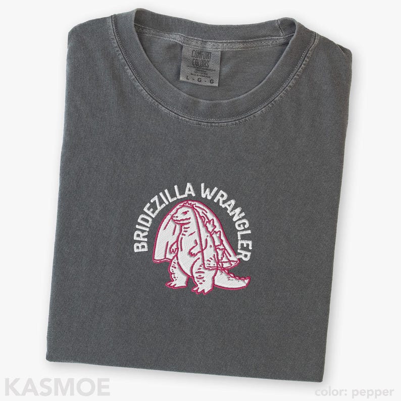 Może przedstawiać: Ciemnoszary t-shirt z wyhaftowaną grafiką przedstawiającą stworzenie przypominające dinozaura w welonie ślubnym. Napis BRIDEZILLA WRANGLER jest wygięty w łuk nad stworzeniem białymi wielkimi literami. Koszulka posiada okrągły dekolt.