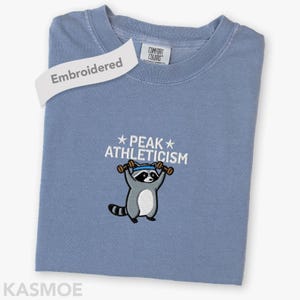 May include: A blue crew neck t-shirt featuring an embroidered graphic of a raccoon wearing a blue headband and lifting two brown dumbbells. Above the raccoon, the white text reads PEAK ATHLETICISM with a star on each side of the word PEAK.