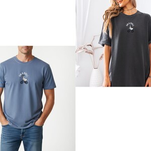 May include: A blue t-shirt and a charcoal gray t-shirt, each featuring a small graphic on the chest of a white goose riding a bicycle with the text "ON A ROLL" arched above the goose in a circular white font.