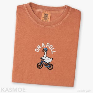 May include: A yam-colored crew neck t-shirt featuring an embroidered graphic of a white goose wearing a blue helmet and riding a blue bicycle. The text ON A ROLL is embroidered in white above the goose. The shirt has a Comfort Colors brand label.