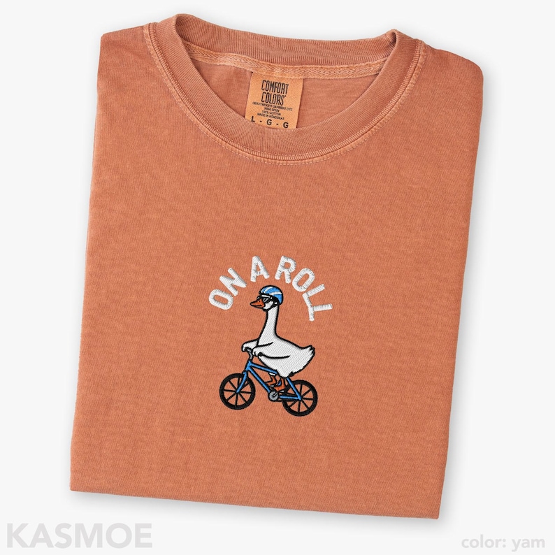 May include: A yam-colored crew neck t-shirt featuring an embroidered graphic of a white goose wearing a blue helmet and riding a blue bicycle. The text ON A ROLL is embroidered in white above the goose. The shirt has a Comfort Colors brand label.