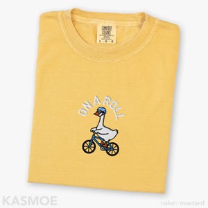 May include: A mustard yellow crew neck t-shirt featuring an embroidered graphic of a white goose wearing a blue helmet and riding a blue bicycle. Above the goose, the text ON A ROLL is embroidered in white. The shirt has a Comfort Colors label.