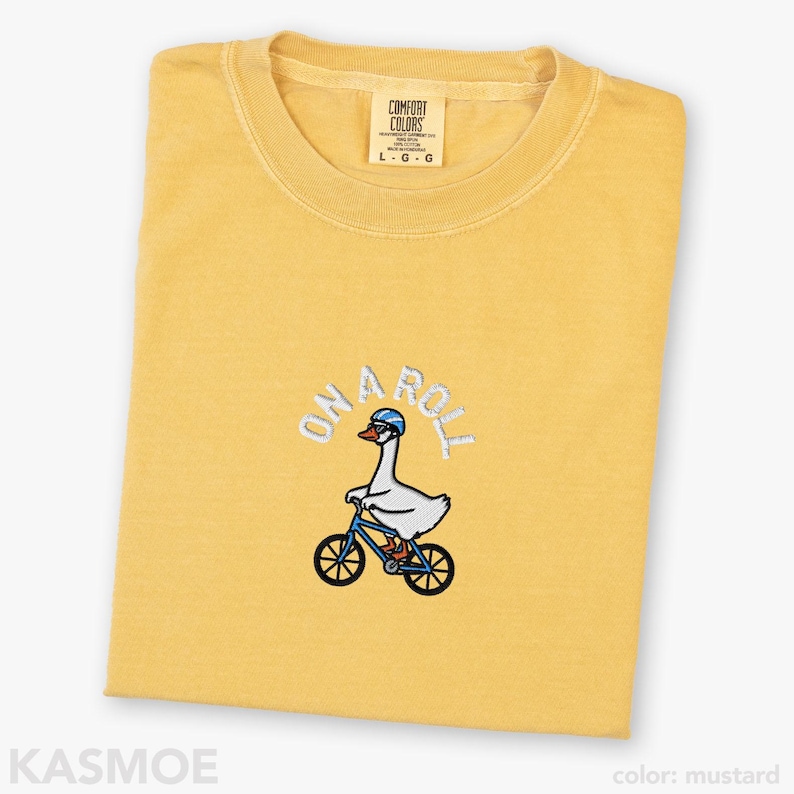 May include: A mustard yellow crew neck t-shirt featuring an embroidered graphic of a white goose wearing a blue helmet and riding a blue bicycle. Above the goose, the text ON A ROLL is embroidered in white. The shirt has a Comfort Colors label.