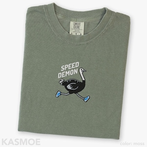 May include: A moss green crew neck t-shirt featuring an embroidered graphic of a black ostrich wearing blue sneakers. Above the ostrich, the text SPEED DEMON is embroidered in white. The shirt has a Comfort Colors brand label at the inner neckline.