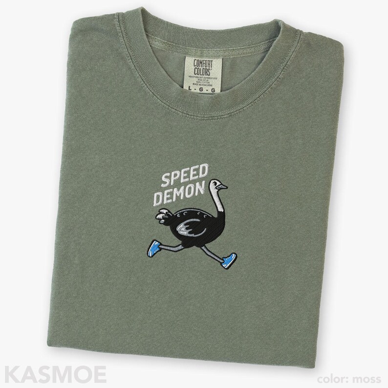 May include: A moss green crew neck t-shirt featuring an embroidered graphic of a black ostrich wearing blue sneakers. Above the ostrich, the text SPEED DEMON is embroidered in white. The shirt has a Comfort Colors brand label at the inner neckline.
