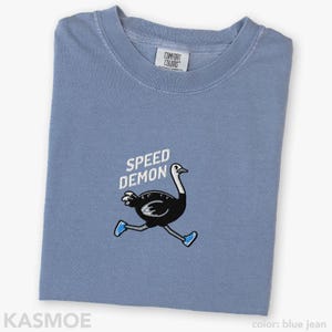 May include: A blue jean colored t-shirt featuring an embroidered graphic of a black ostrich running while wearing blue sneakers. Above the ostrich, the text SPEED DEMON is embroidered in white. The shirt has a Comfort Colors brand neck label.