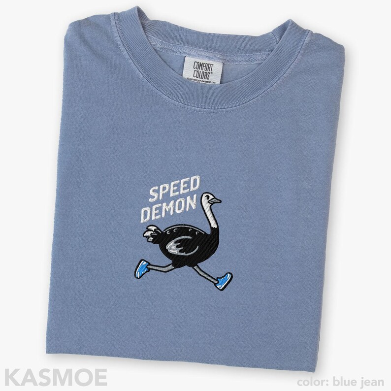 May include: A blue jean colored t-shirt featuring an embroidered graphic of a black ostrich running while wearing blue sneakers. Above the ostrich, the text SPEED DEMON is embroidered in white. The shirt has a Comfort Colors brand neck label.