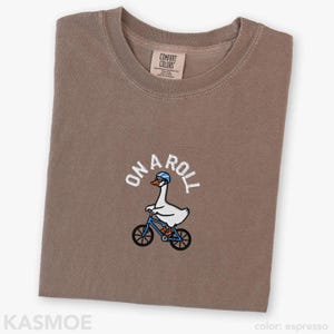 May include: An espresso-colored t-shirt featuring an embroidered graphic of a white goose wearing a blue helmet and glasses while riding a blue bicycle. The text ON A ROLL is embroidered in white above the goose. The shirt has a crew neckline.