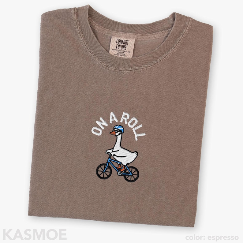 May include: An espresso-colored t-shirt featuring an embroidered graphic of a white goose wearing a blue helmet and glasses while riding a blue bicycle. The text ON A ROLL is embroidered in white above the goose. The shirt has a crew neckline.