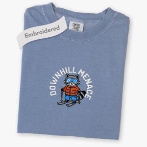 May include: A blue crewneck t-shirt featuring an embroidered graphic of a raccoon wearing a red jacket, blue pants, and blue ski goggles while skiing. The text "DOWNHILL MENACE" is arched above the raccoon. A white tag reads "Embroidered".