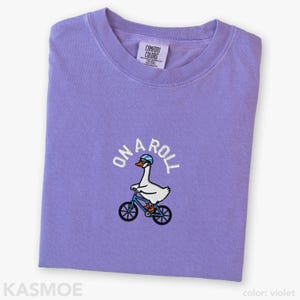 May include: A violet crewneck t-shirt featuring an embroidered graphic of a white goose wearing a blue helmet and riding a blue bicycle. Above the goose, the text ON A ROLL is embroidered in white. The shirt has a Comfort Colors brand label.