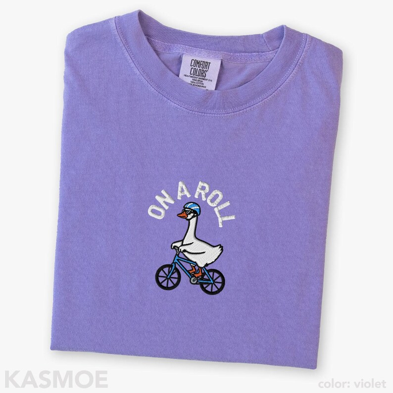 May include: A violet crewneck t-shirt featuring an embroidered graphic of a white goose wearing a blue helmet and riding a blue bicycle. Above the goose, the text ON A ROLL is embroidered in white. The shirt has a Comfort Colors brand label.
