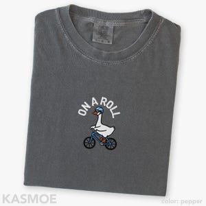 May include: A charcoal gray crew neck t-shirt featuring an embroidered graphic of a white goose wearing a blue helmet and riding a blue bicycle. The text ON A ROLL is arched above the goose in white lettering. The shirt has a Comfort Colors label.
