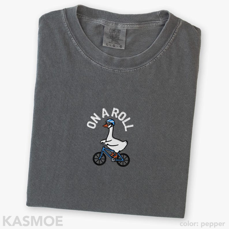 May include: A charcoal gray crew neck t-shirt featuring an embroidered graphic of a white goose wearing a blue helmet and riding a blue bicycle. The text ON A ROLL is arched above the goose in white lettering. The shirt has a Comfort Colors label.