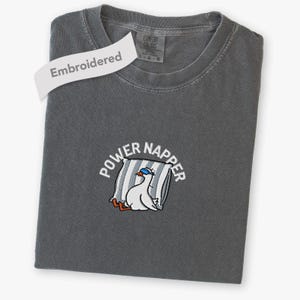 May include: A charcoal grey crew neck t-shirt featuring an embroidered graphic of a white goose wearing blue sleep goggles, resting against a striped pillow. The text "POWER NAPPER" is arched above the goose in white, and a tag reads "Embroidered".