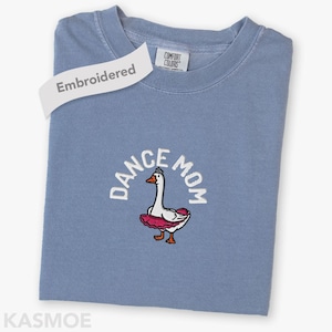 May include: A blue crewneck t-shirt featuring an embroidered graphic of a white goose wearing a pink tutu and a small silver crown. The text "DANCE MOM" is arched above the goose in white thread. The shirt has a Comfort Colors brand label.