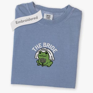May include: A blue short-sleeved t-shirt featuring an embroidered graphic of a green frog wearing a white bridal veil and holding a bouquet of white flowers. The text "THE BRIDE" is embroidered in white above the frog. The shirt has a Comfort Colors label.