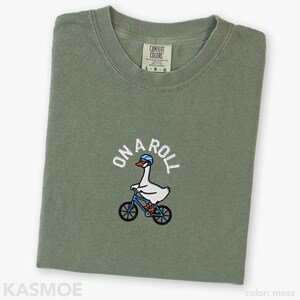 May include: A moss green t-shirt featuring an embroidered graphic of a white goose wearing a blue helmet and glasses while riding a blue bicycle. The text "ON A ROLL" is arched above the goose in white lettering. The shirt has a Comfort Colors label.