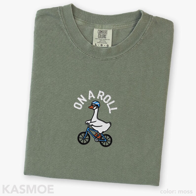 May include: A moss green t-shirt featuring an embroidered graphic of a white goose wearing a blue helmet and glasses while riding a blue bicycle. The text "ON A ROLL" is arched above the goose in white lettering. The shirt has a Comfort Colors label.