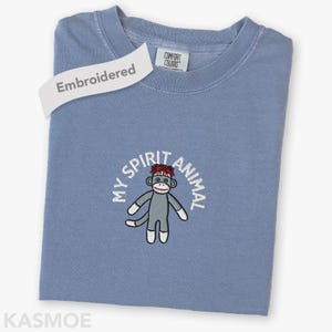 以下が含まれることがあります： 刺繍された靴下猿のデザインが施された水色のTシャツ。「MY SPIRIT ANIMAL」というフレーズが猿の周りに刺繍されています。シャツには「Comfort Colors」のタグと「Embroidered」と書かれたラベルが付いています。