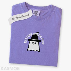 May include: A light purple crewneck t-shirt featuring an embroidered graphic of a white ghost wearing a black witch hat. The text "RESTING WITCH FACE" is embroidered in white above the ghost. A white tag reads "Embroidered" and "KASMOE".